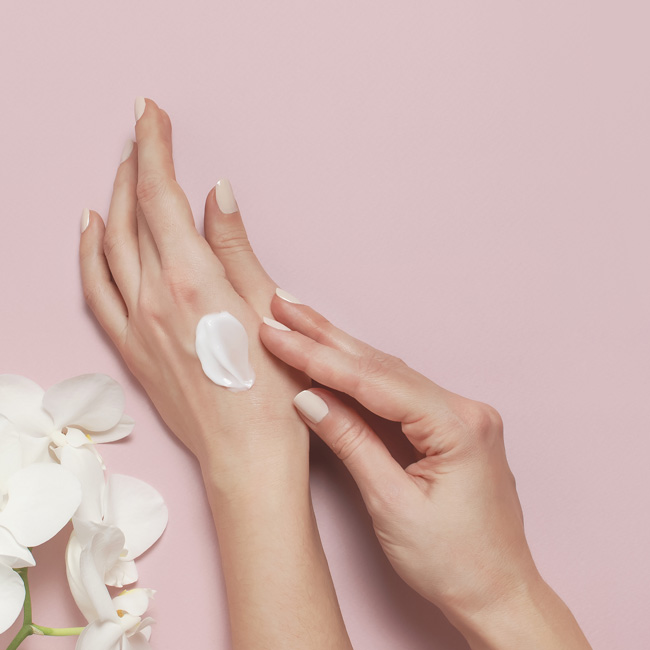 Young woman moisturizes her hand with cosmetic cream lotion opened container with cream body milk White Phalaenopsis orchid flowers on pink background Flat lay top view minimalism style Beauty concept